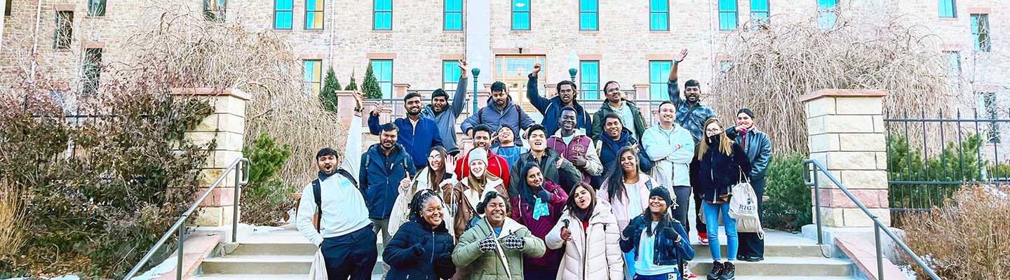  a large group of international students smile and wave as they pose for the camera on the steps on Main Hall on the Regis campus